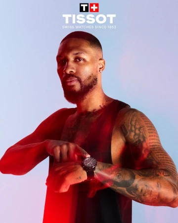 Promotional shot of an adult male model pointing to a Tissot watch on his wrist against a light blue backdrop with red lighting accents.