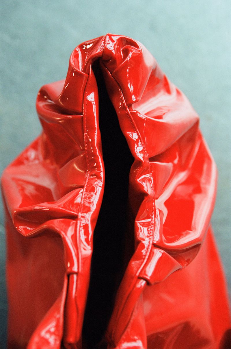 Close-up of a shiny, bright red vinyl-like material bunched and gathered at the top against a grey background, creating deep shadows.