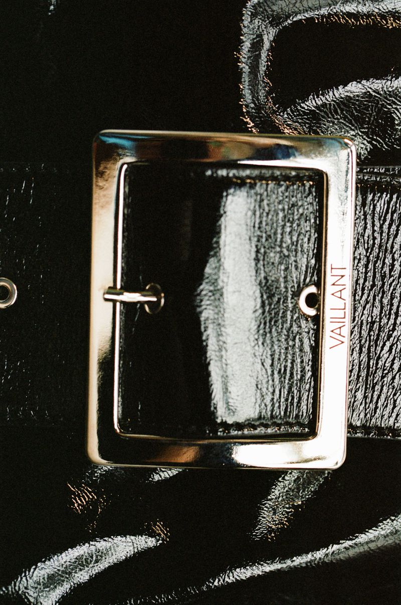 Close-up on shiny black vinyl belt with a metal buckle. The buckle is square with rounded corners, and the brand name 'Vaillant' is visible.