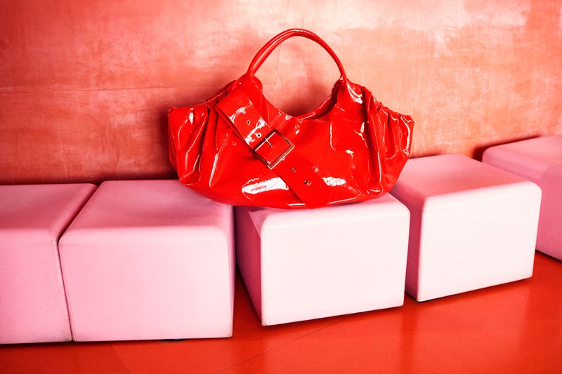 Red patent leather purse with buckle detail displayed on pink cushioned cubes against a red textured wall and glossy red floor.