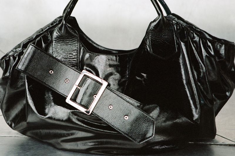 Close-up of a stylish black patent leather hobo bag featuring a wide belt detail with a silver buckle. Fashionable accessory.