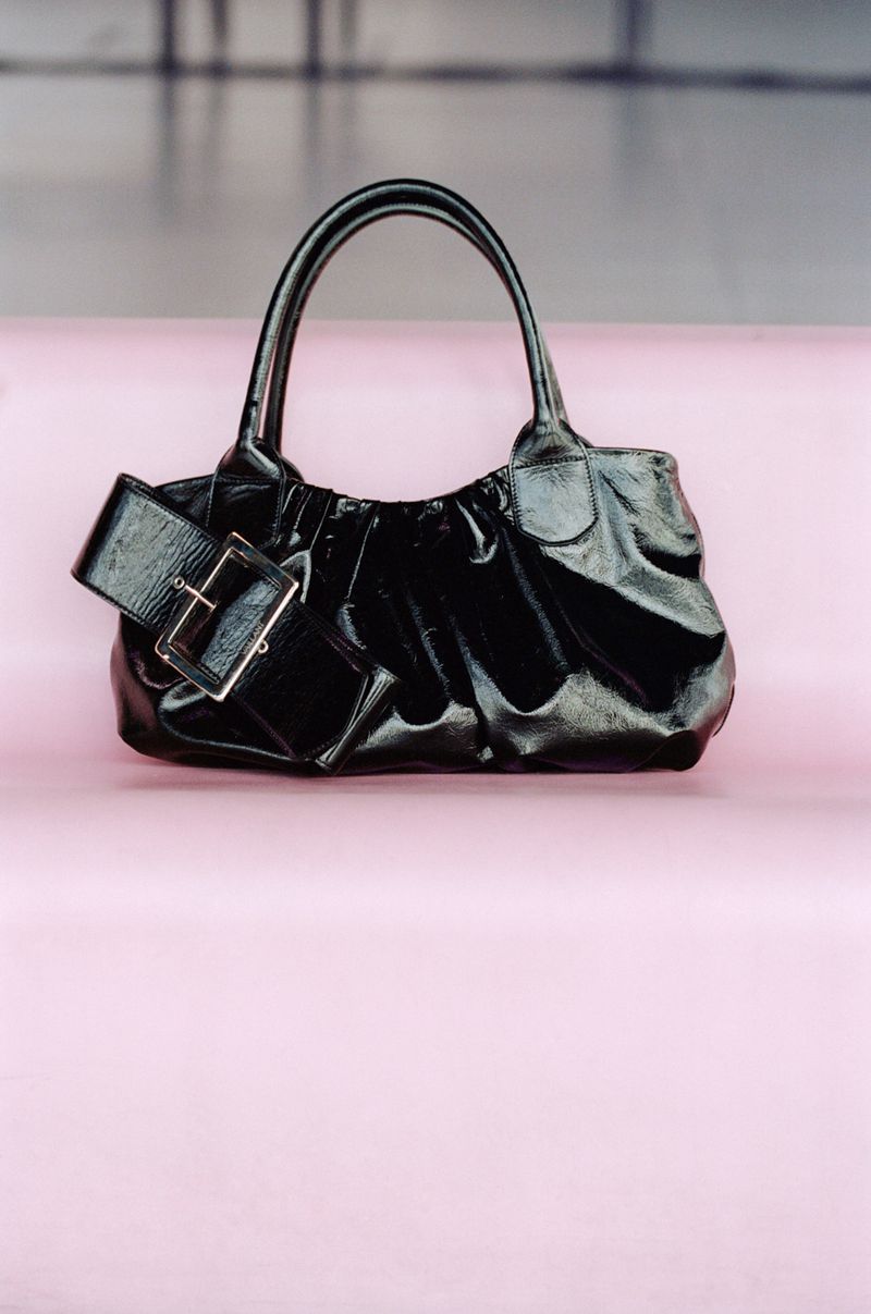 Elegant black patent leather purse with a buckle strap on a pink surface against a blurred gray and white background.