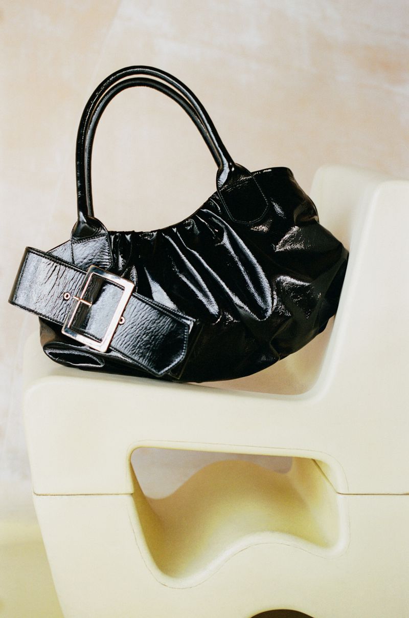 Shiny black patent leather purse with a wide buckle detail on a cream-colored chair in a studio with a neutral background.