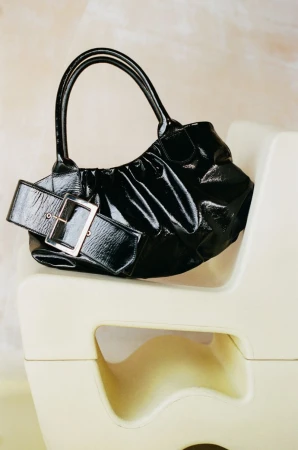Black patent leather ruched handbag with large silver buckle strap resting on a cream sculptural chair.