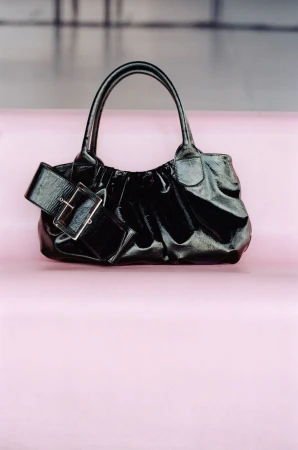 Vaillant black glossy gathered leather handbag with oversized buckle strap detail displayed on a pale pink surface.