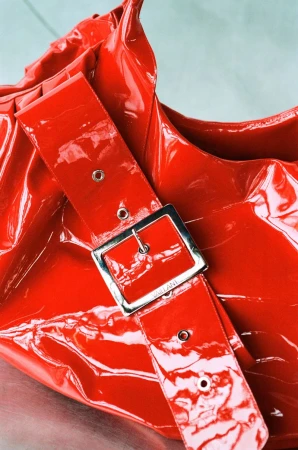 Close-up of a glossy red patent leather Vaillant handbag with a wide strap and large silver metal buckle.