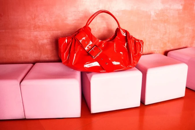 Red patent leather handbag with a wide buckle strap resting on pink modular cube seats against a textured red wall.