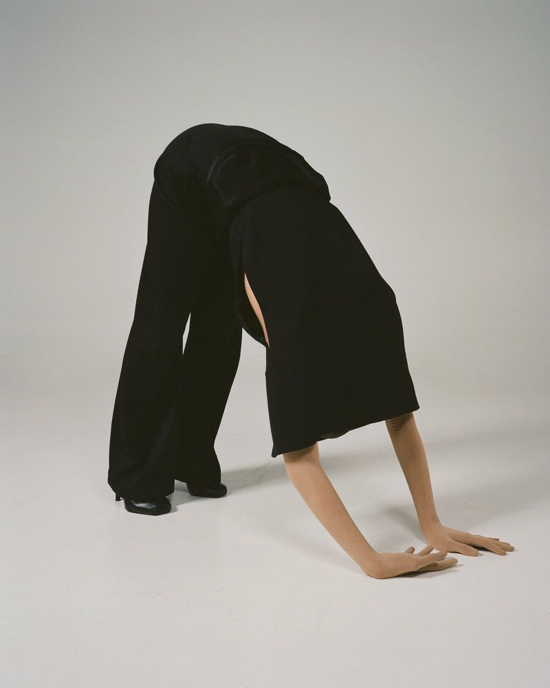 Fashion image of a flexible person in black clothing bending over and placing their hands on a white floor, showcasing body contortion.