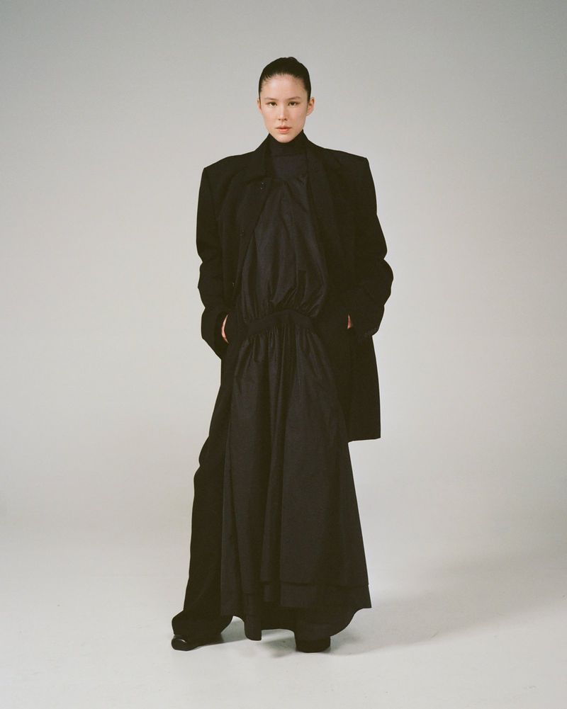 Fashion portrait of an adult female wearing a long, dark dress and coat in a studio. She poses with a neutral expression.