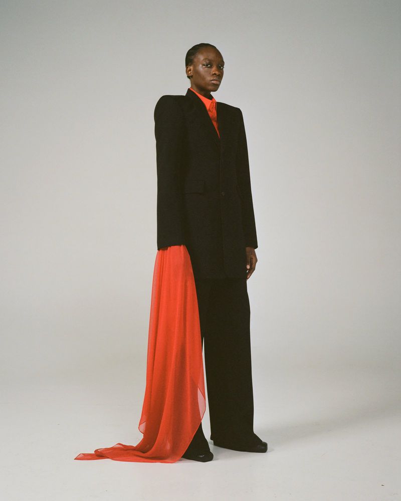 An adult woman in a black suit and red scarf poses in a photo studio with a full shot on a white background. Fashion editorial.