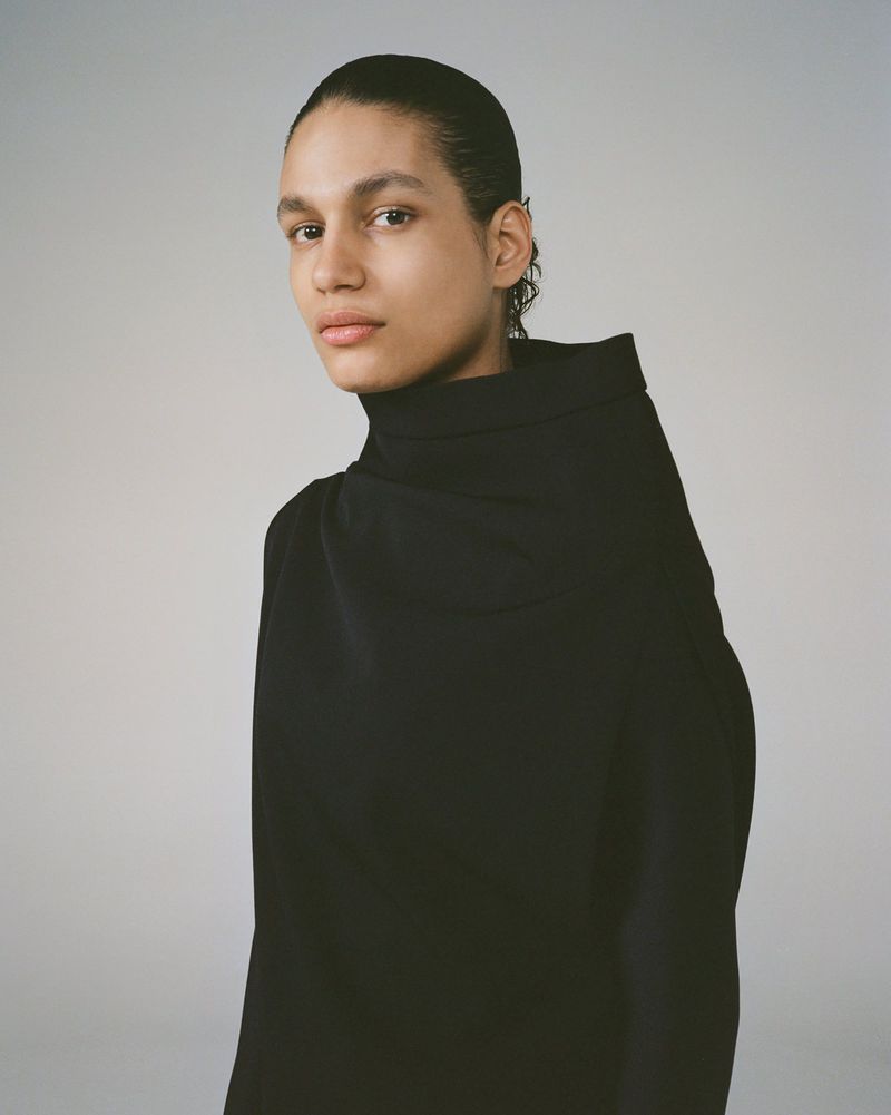 Portrait of a woman with slicked-back hair in a high-necked black dress, posing in front of a pale gray background.