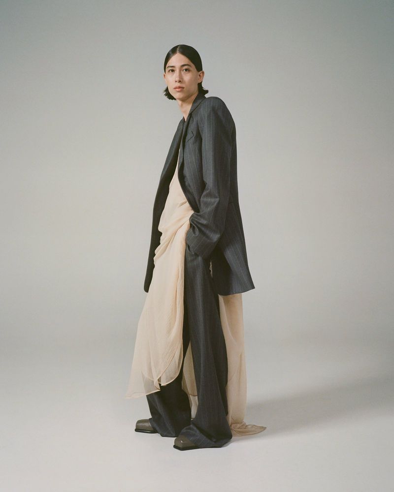 A person poses in a dark suit jacket and wide-leg pants, layered over a flowy, draped top, in a fashion studio shot on a neutral background.