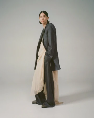A person poses in a dark suit jacket and wide-leg pants, layered over a flowy, draped top, in a fashion studio shot on a neutral background.
