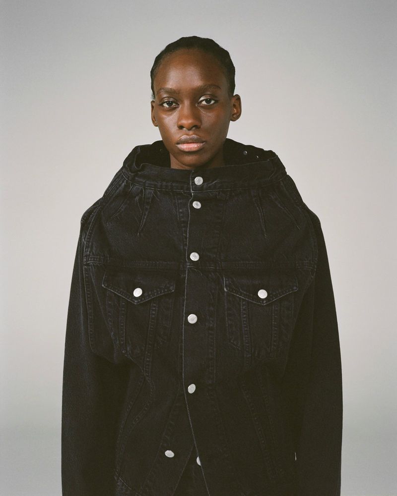 Portrait of a dark-skinned female fashion model wearing a dark denim jacket with metal button closures against a light grey backdrop.