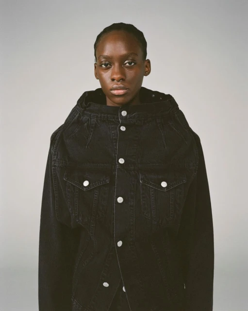 Portrait of a dark-skinned female fashion model wearing a dark denim jacket with metal button closures against a light grey backdrop.