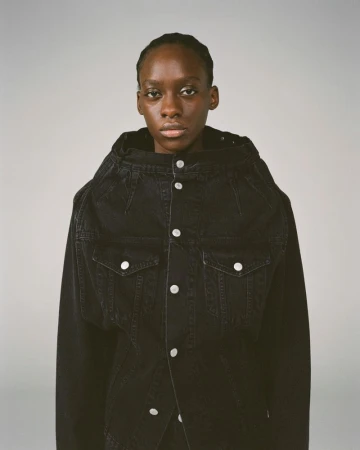 Portrait of a dark-skinned female fashion model wearing a dark denim jacket with metal button closures against a light grey backdrop.