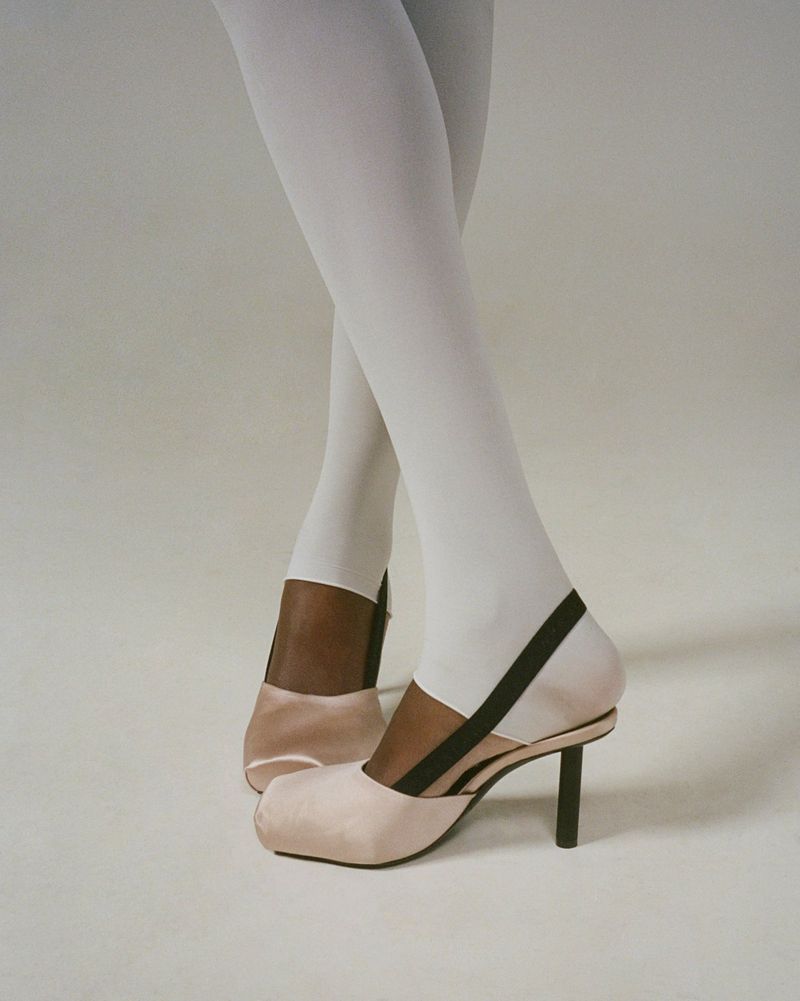 Close-up of legs wearing white stirrup tights and blush pink satin slingback heels with black straps on a pale seamless studio floor.