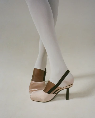 Close-up of legs wearing white stirrup tights and blush pink satin slingback heels with black straps on a pale seamless studio floor.