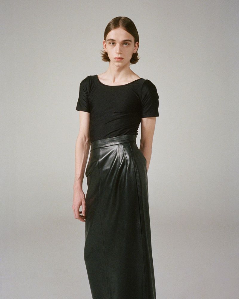 Person poses in a black shirt and long leather skirt against a grey backdrop, fashion shoot, studio lighting.