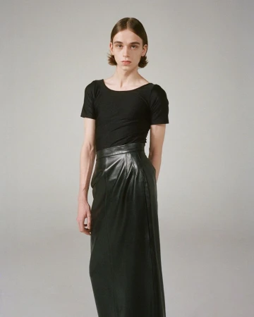 Person poses in a black shirt and long leather skirt against a grey backdrop, fashion shoot, studio lighting.