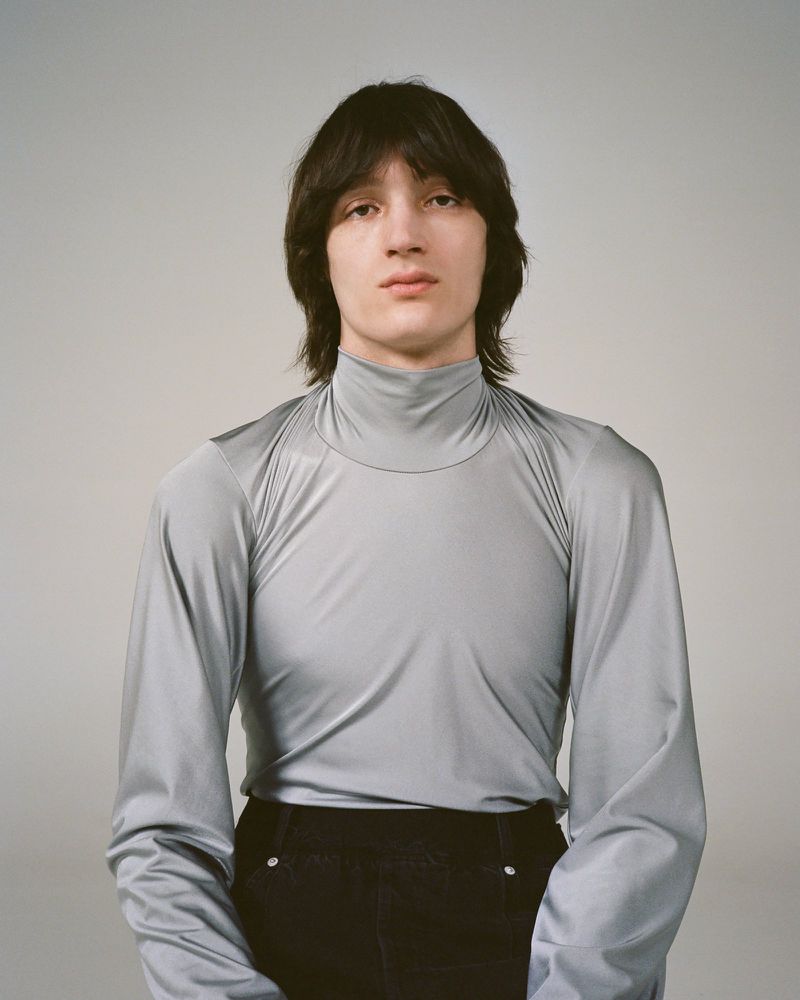 Portrait of a young adult with dark hair wearing a grey turtleneck and black pants in a photo studio setting on a neutral background.