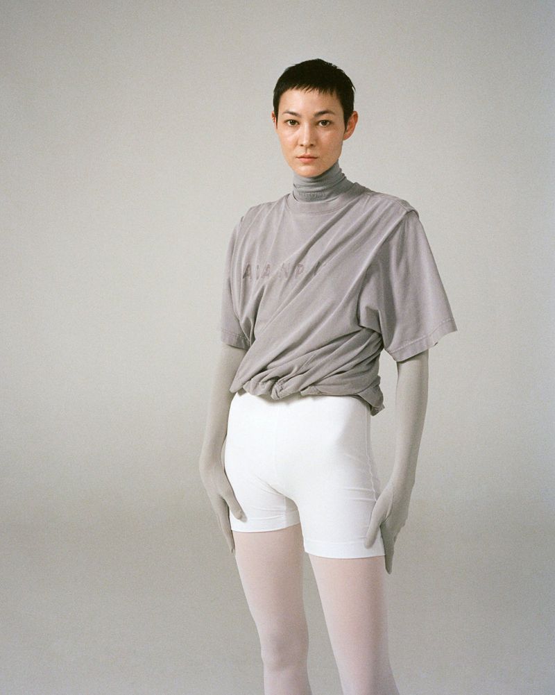 Woman in gray top and white shorts standing in photo studio. Female with short hair posing. Light gray background. Portrait orientation.