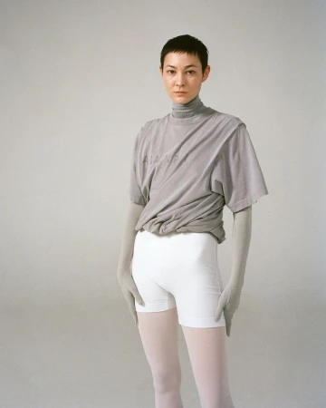 Woman in gray top and white shorts standing in photo studio. Female with short hair posing. Light gray background. Portrait orientation.
