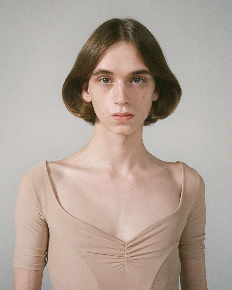Portrait of a light-skinned adult with shoulder-length brown hair, wearing a beige, long-sleeved top, standing against a grey background.