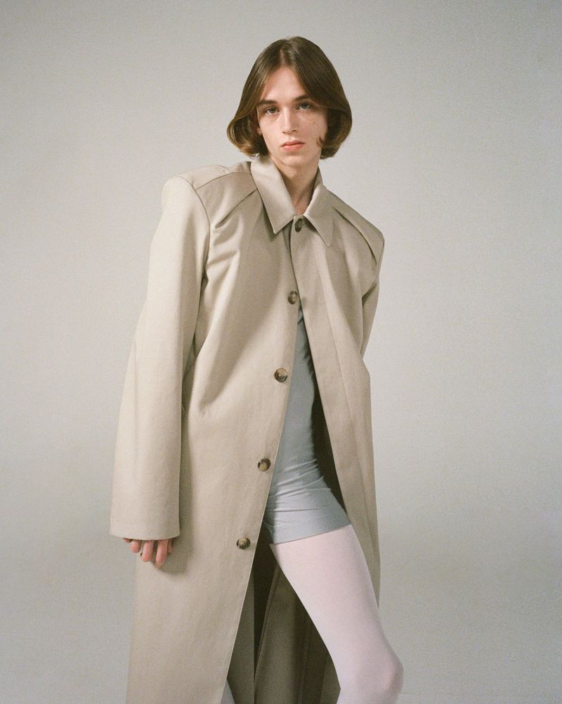 Model with bobbed brown hair posing in an oversized beige trench coat, grey bike shorts, and pale tights against a neutral grey background.