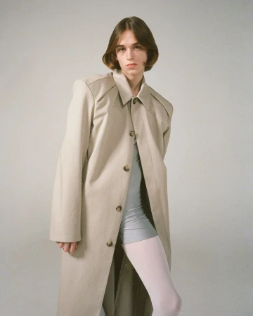 Model with bobbed brown hair posing in an oversized beige trench coat, grey bike shorts, and pale tights against a neutral grey background.