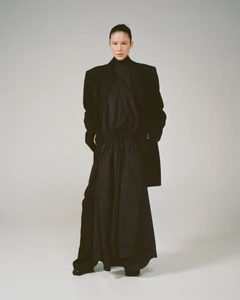 Model standing against a plain light grey backdrop wearing an oversized black coat over a dark dress.