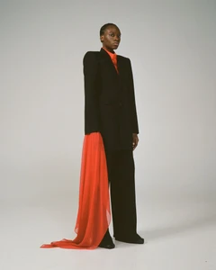 Model in a black suit with structured shoulders and a long red fabric hanging from the sleeve against a grey studio background
