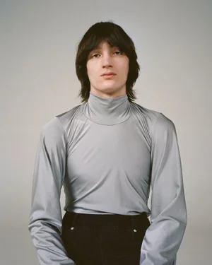 Model wearing a light grey long-sleeve turtleneck top and dark trousers against a neutral studio background.