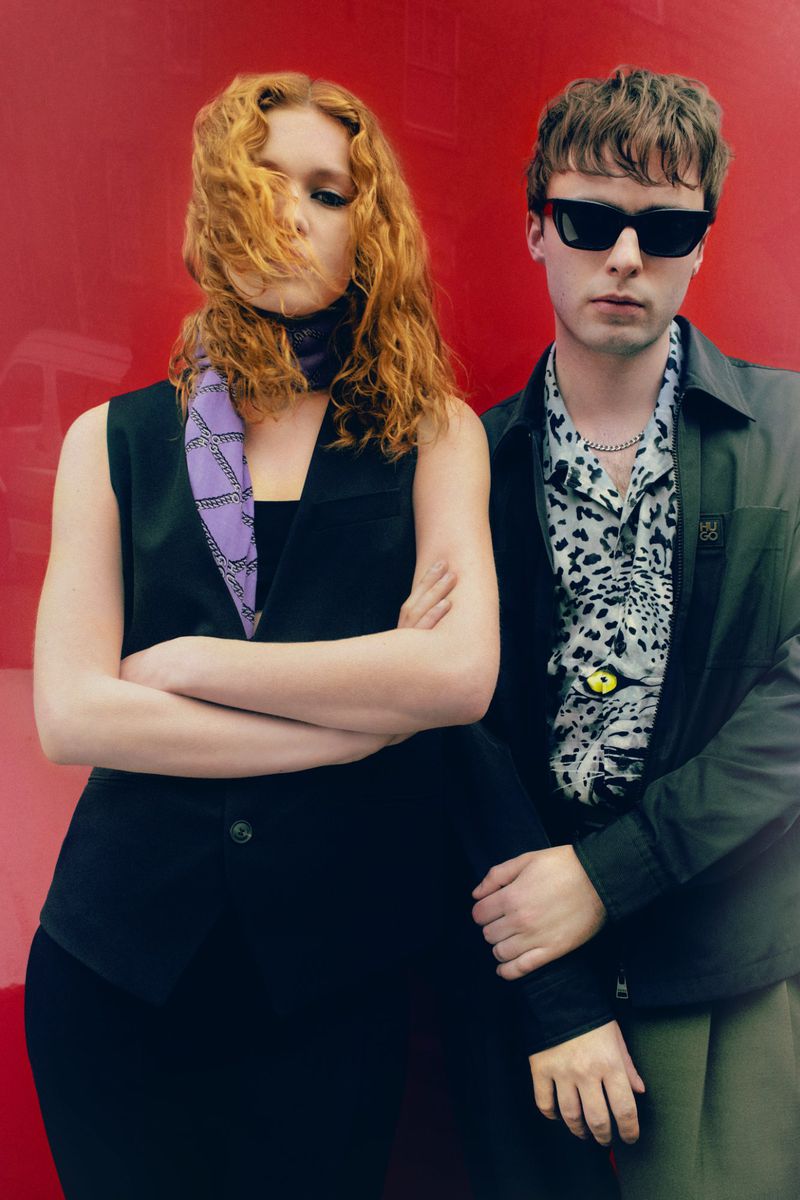 Two people pose against a solid red backdrop. A red-haired woman with a scarf and a man in sunglasses with a patterned shirt.