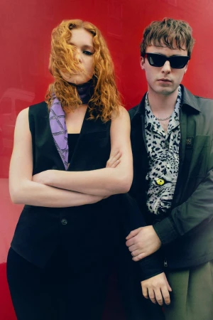 Two people pose against a solid red backdrop. A red-haired woman with a scarf and a man in sunglasses with a patterned shirt.