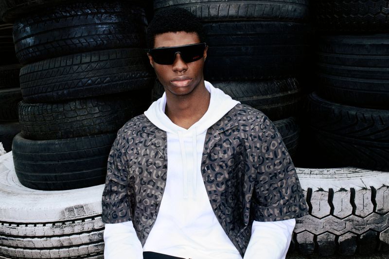Young adult poses with sunglasses, wearing patterned jacket and white hoodie, sitting against backdrop of stacked black tires.