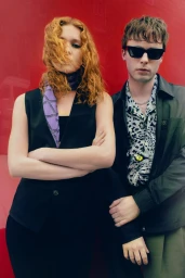 Two models posing against a bold red background, featuring a woman with curly red hair and a man in sunglasses.