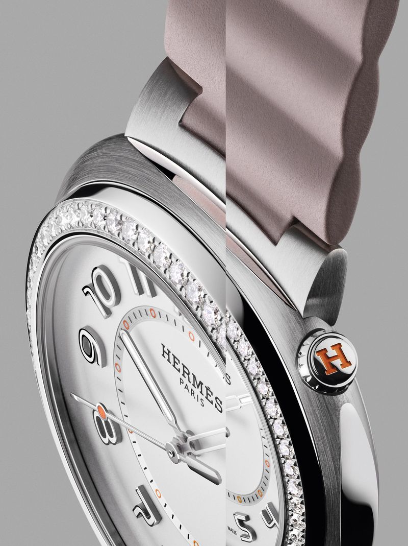 Close-up of a silver Hermès watch with a diamond bezel, a white dial with Arabic numerals, and a light pink watch band, against a gray background.