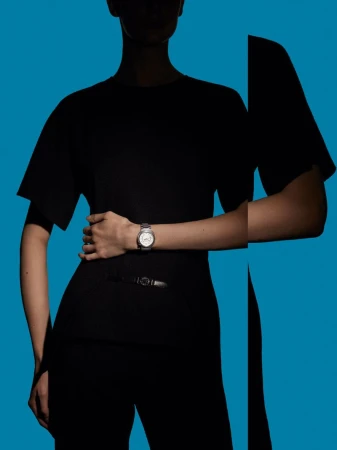 A woman in a black outfit wears a silver wristwatch against a blue background in a fashion shot emphasizing wristwear and minimalistic style.