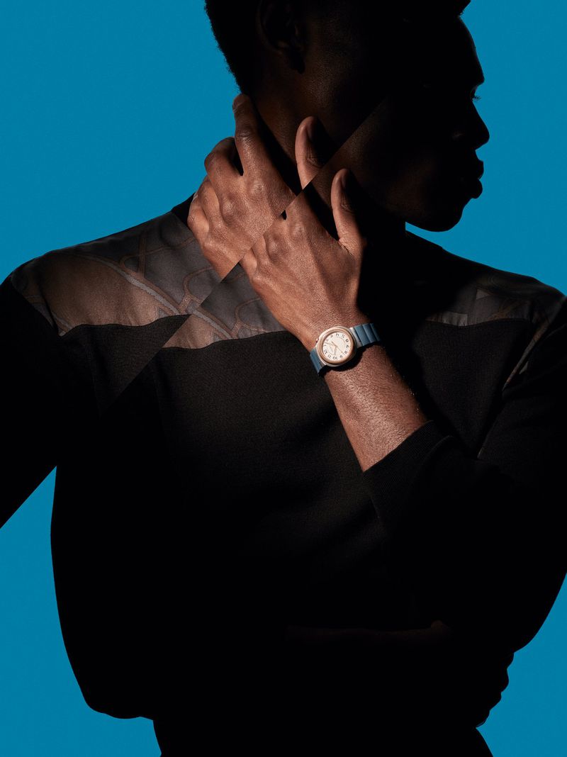 Close-up studio shot of an adult with dark skin wearing a black shirt and wrist watch, hands around neck, against a blue background.