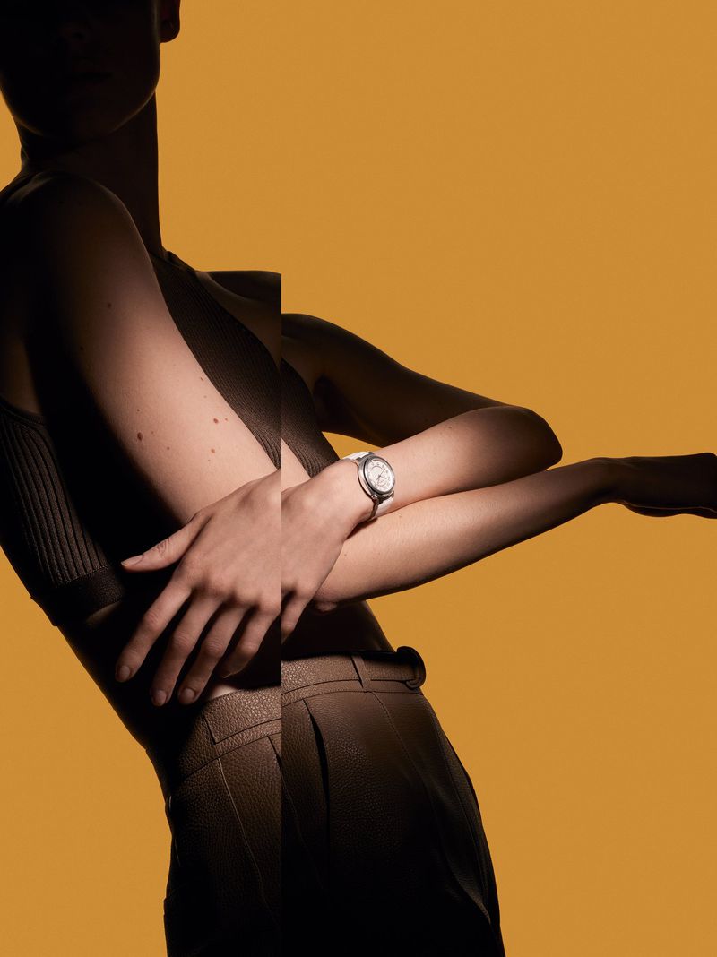 Stylish wristwatch on a slender, lithe woman against a yellow backdrop, partially veiled in shadows for dramatic effect.