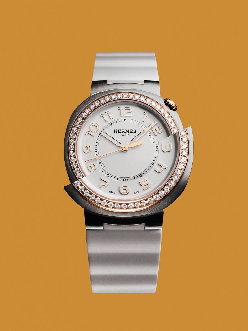 Elegant silver Hermès watch with a round face, diamond bezel, and silver band on a mustard-yellow background, showcasing luxury and style.