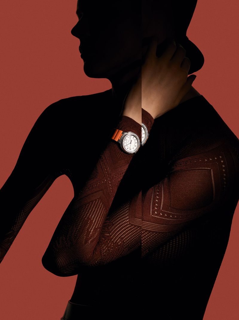 Close-up studio shot of a person with a fashionable watch, a textured top, and a hat against a warm red backdrop.