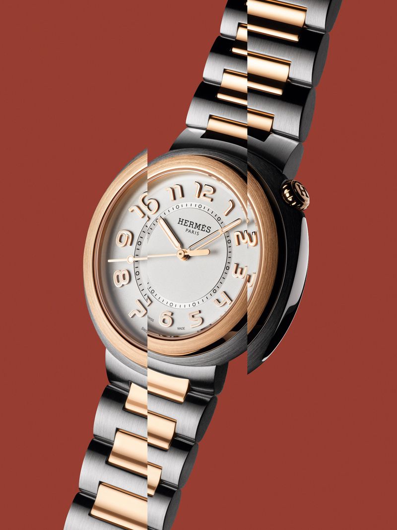 Silver and gold Hermès watch with a round white face and a metal band, set against a reddish-brown background in a studio shot.