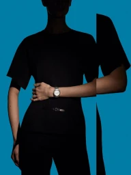 Model wearing a black short-sleeved top and silver luxury wristwatch with hand on hip against a vivid blue background.