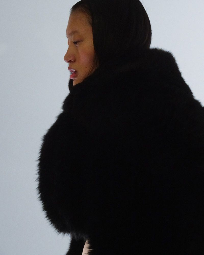 Profile of a female fashion model in a large dark fur coat with a hood, against a gradient background.