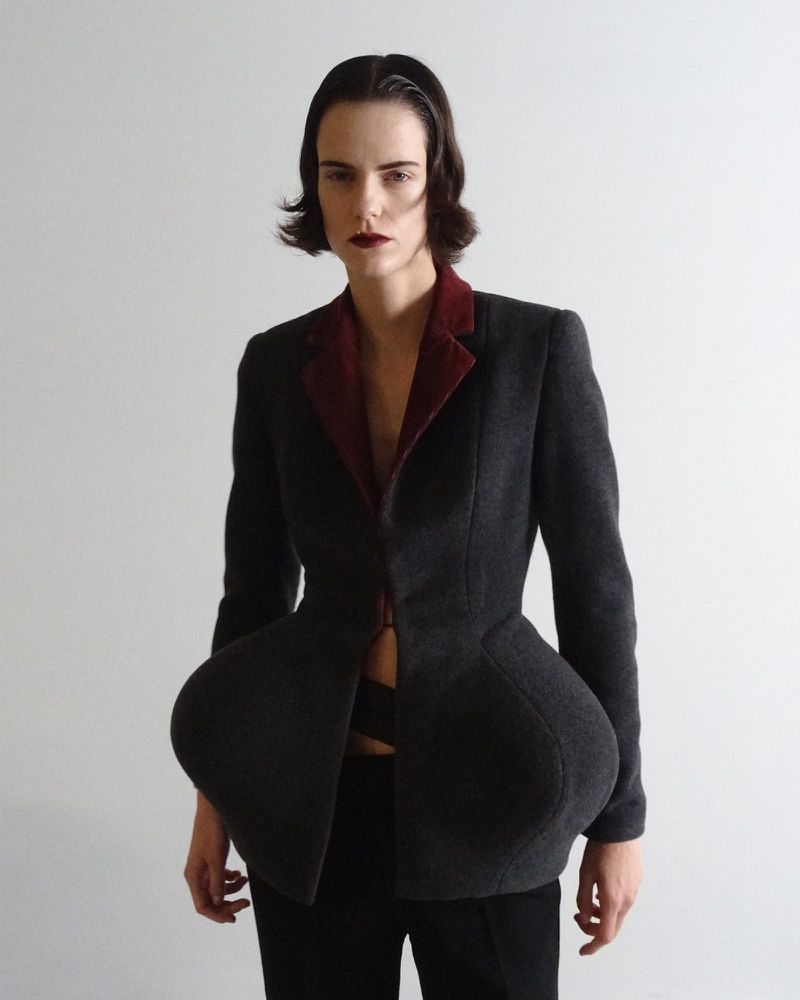 A person is posing in a gray blazer jacket with burgundy lining and black pants against a white backdrop in a studio environment.