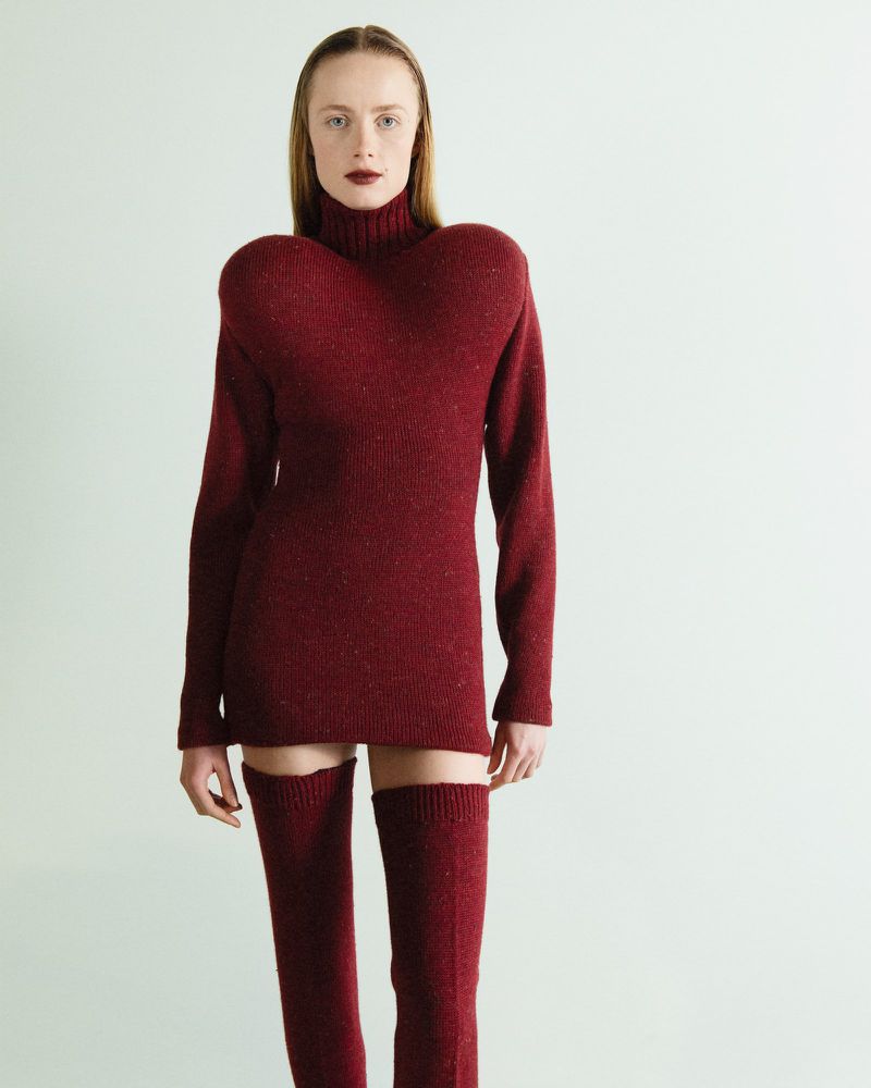 A female model in a burgundy knit turtleneck dress and thigh-high socks poses against a light background in a fashion forward studio shot.