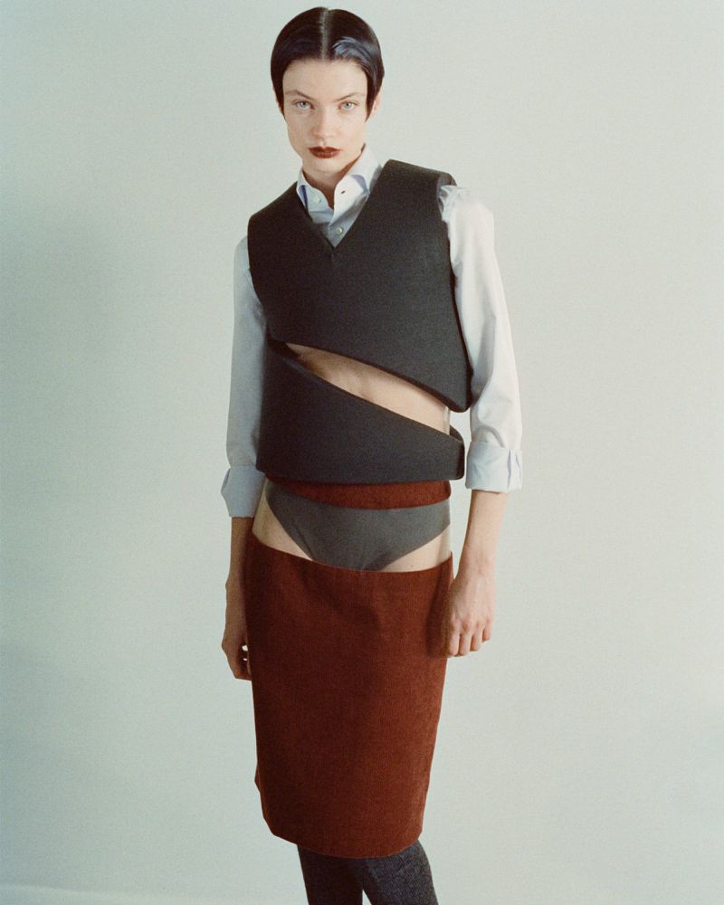 Fashion editorial shot of a woman with short hair, wearing a white collared shirt and deconstructed vest and skirt with leg coverings.