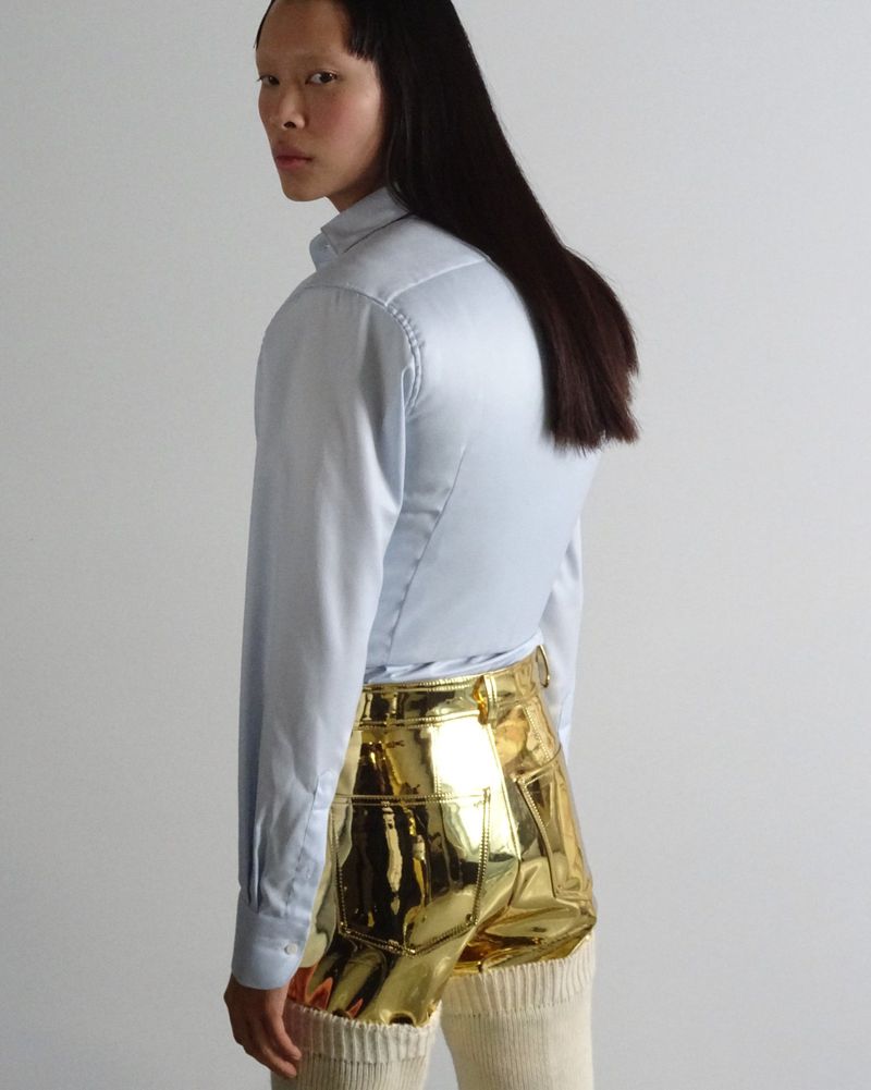 A woman in a light blue button-down shirt and gold shorts poses against a white background, looking over her shoulder.